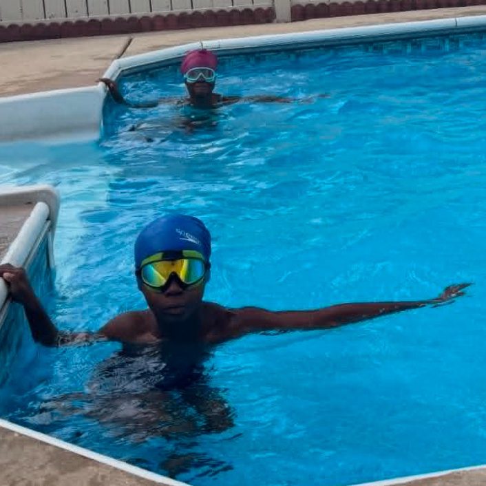 Two children in the pool, wearing swim caps and goggles with one hand on the wall ready to swim.